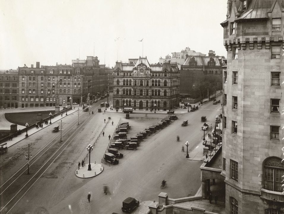 A sepia photograph from the fourth floor of a building, overlooking a triangular public plaza, many old cars with canvas tops are parked in the square. Neo-gothic buildings make up 2 borders of the square, and a set of tram tracks comprise the third