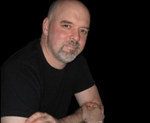 A bald man wearing a black t-shirt looks at the camera while sitting against a dark background.
