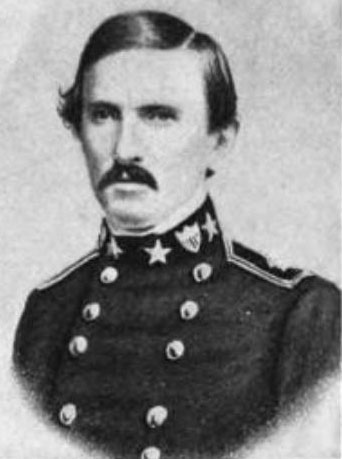 A man in his early forties with short black hair and a mustache. He is wearing a black military coat with two rows of buttons down the front and various military insignia on the collar.