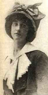 A white woman wearing a large hat and a suit with a complicated white collar or scarf.