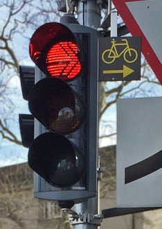 A traffic signal with a small permanent sign next to the red light. The sign shows a yellow bicycle above a yellow arrow pointing towards the right.