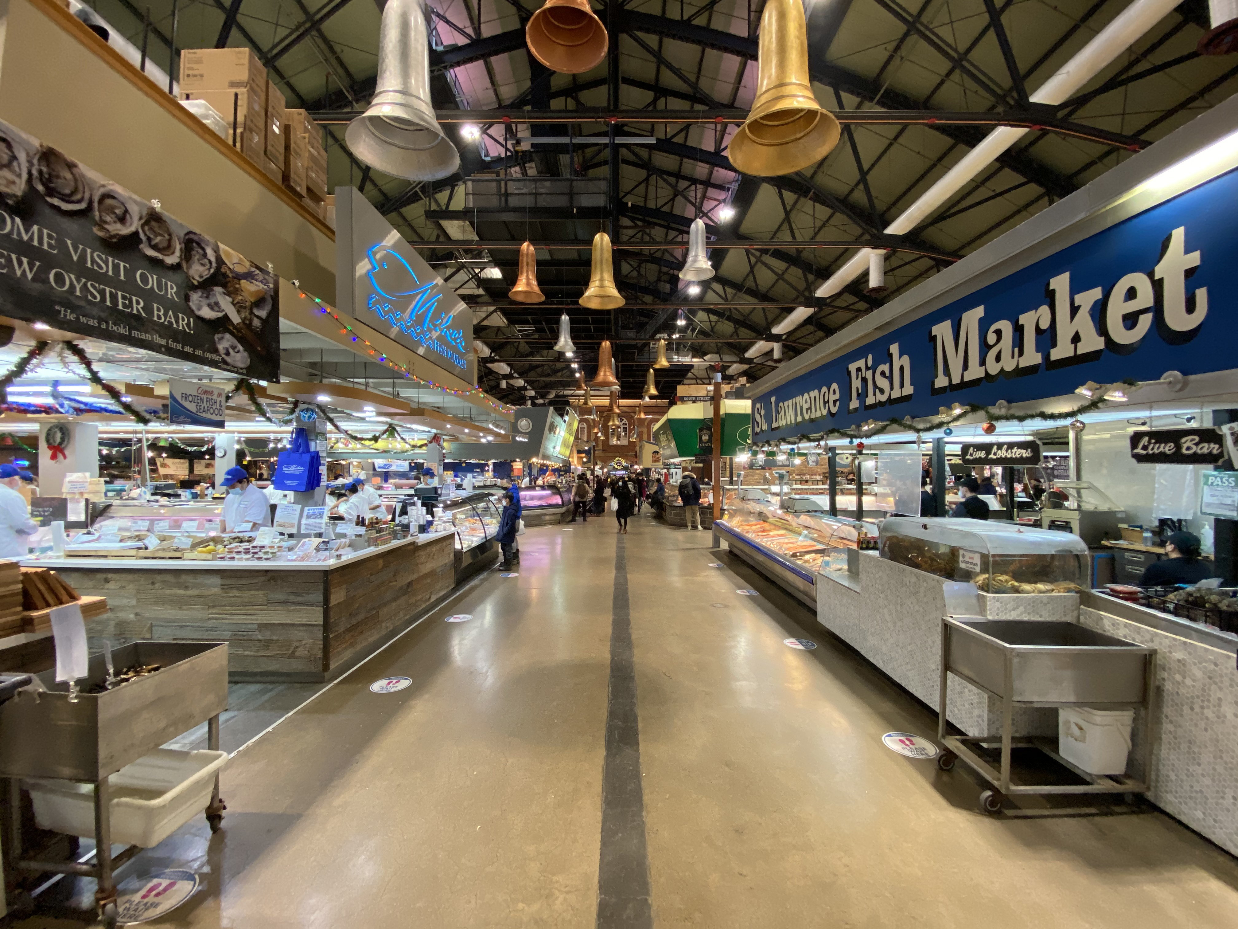 A beige walkway is shown with stores on either side. Oyster and Fish are being sold from the camera’s point of view. Customers can be seen walking in bunches towards the camera, with a brown brick wall behind them.