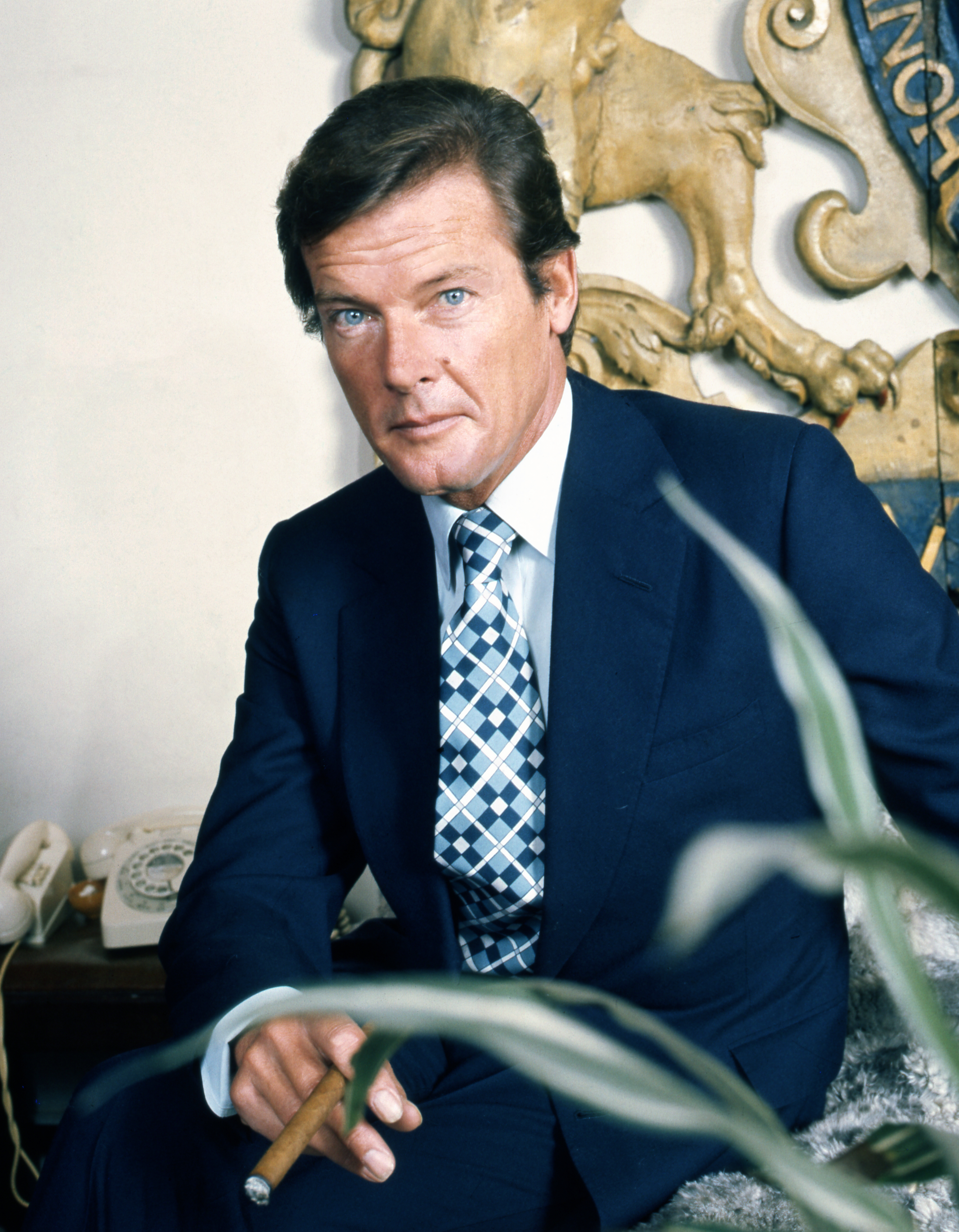 Roger Moore sitzend in einem dunkelblauen Anzug weißen Hemd und einer blau