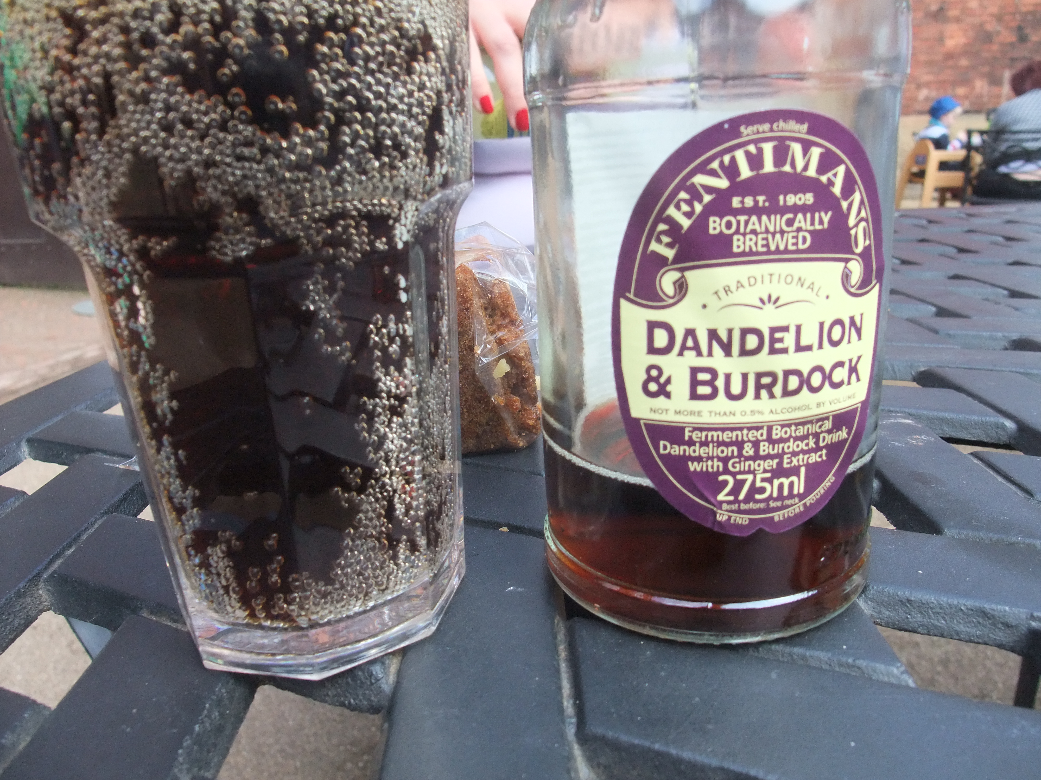 A bottle of dandelion and burdock, which is a dark-coloured fizzy drink, beside a glass of the drink.