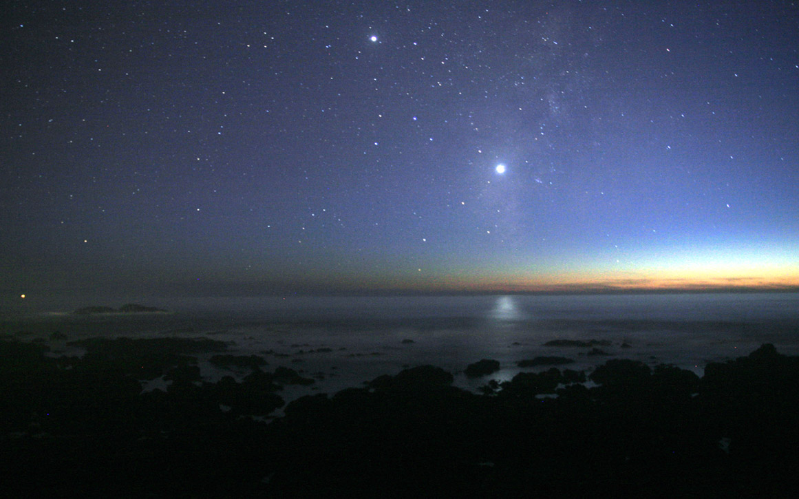 A photograph of the night sky taken from the seashore. A glimmer of sunlight is on the horizon. There are many stars visible. Venus is at the centre, much brighter than any of the stars, and its light can be seen reflected in the ocean.