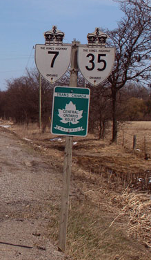 "An image of a signpost on a clear winter day. Behind the signpost are several leafless trees. To the left of the foot of the sign is the gravel shoulder of the road (not pictured) that it stands beside. The signpost is wooden, with three signs. Two are at the top, and one is centred below those. The top two are bullet-shaped signs with a king's crown on top. One is for Highway 7 and one for Highway 35. The sign below is green, with a white maple leaf in the centre. Above the leaf is a white banner with green text, reading "TRANS-CANADA HIGHWAY". The centre of the leaf reads in green "CENTRAL ONTARIO ROUTE". Finally, a white banner with a hung appearance is below the leaf. The green text within the banner reads "ONTARIO""