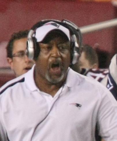 Candid head and shoulders photograph of Fears wearing a white New England Patriots polo shirt and a coaches headset on a football sideline