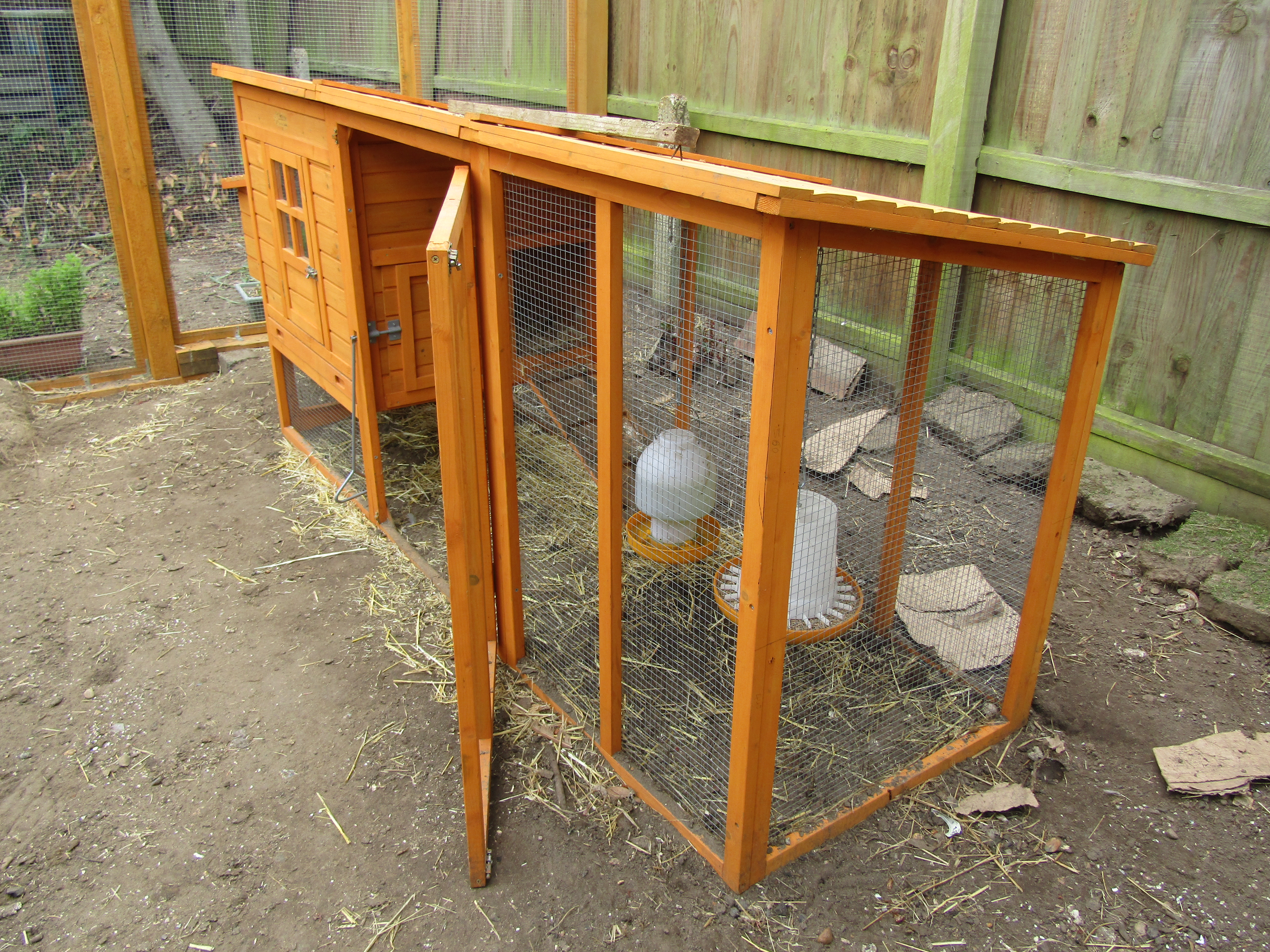 A wooden henhouse with a wood-framed coop with feeders is set within a larger fenced yard