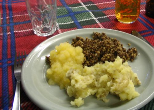 On a plaid table cloth, two cups, one filled with a brown beverage, are featured at the top of the image. A white plate with Scottish foods haggis, neaps, and tatties fills most of the image, with a silver fork and knife on either side. 