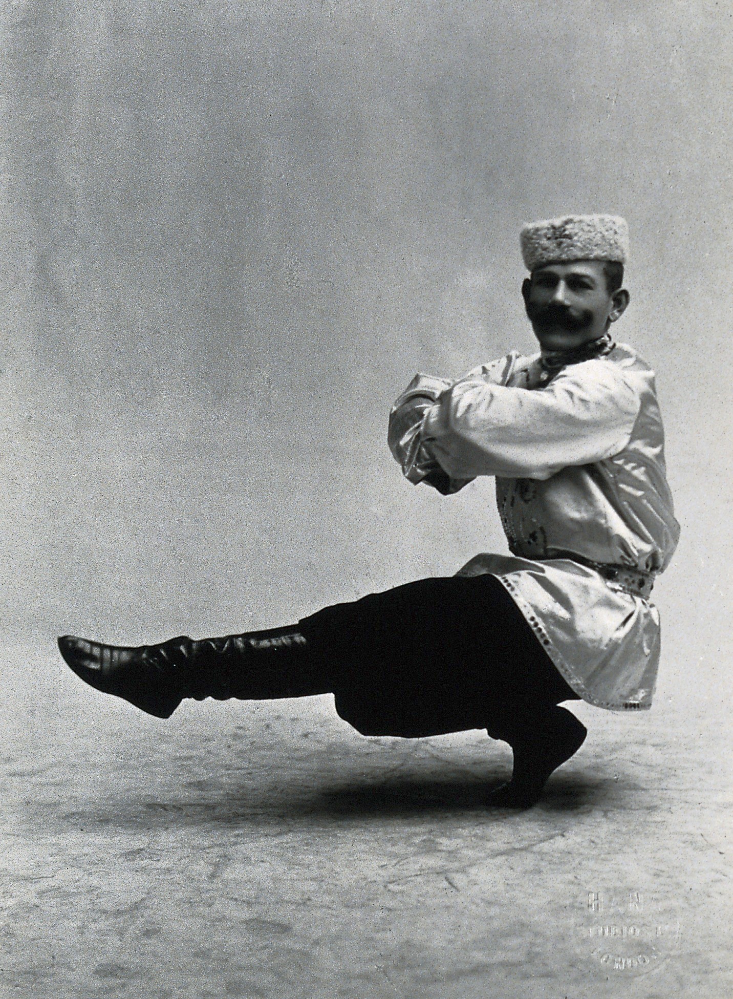 A man, in a satin tunic and fur hat, dancing a "Cossack" measure in a studio setting. Photograph, ca.1899-1908.jpg