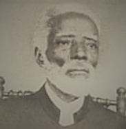 Photographie en noir et blanc et en gros plan d'un homme noir à la barbe blanche, habillé d'un vêtement religieux noir et adossé à une chaise.