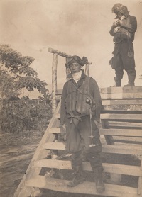 Photo noir et blanc d'un homme en tenue de pilote sur un escalier en bois. À l'arrière-plan, un autre pilote en tenue est debout en haut de l'escalier.