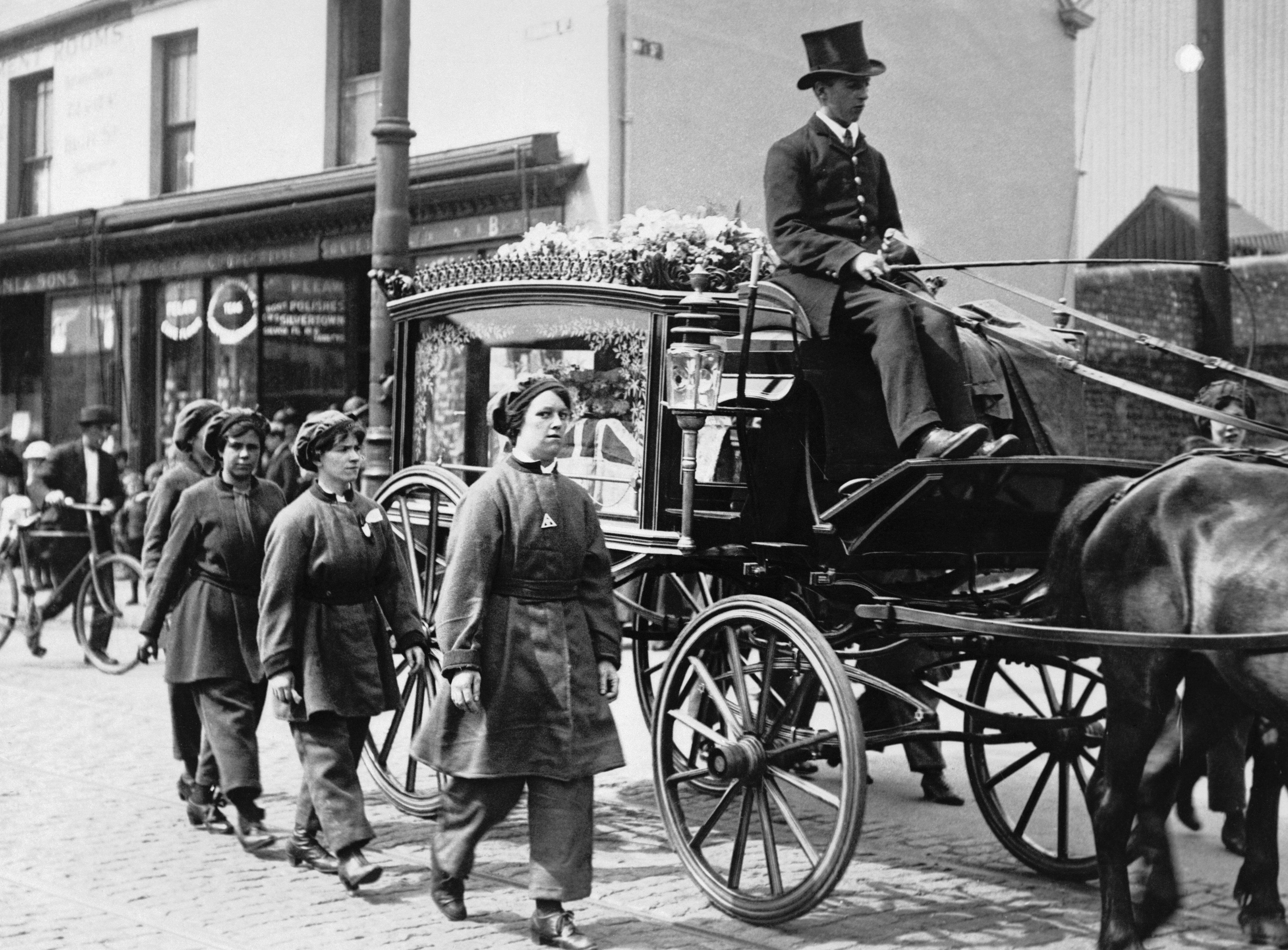 “Female munitions workers walk beside the horsedrawn hearse of one of their colleagues who had been killed at work, August Bank Holiday 1917. They are wearing their factory uniforms as a sign of respect during the funeral procession through Swansea.“