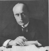 Fotografie alb-negru a bustului unui bărbat cu chelie avansată, în costum, cravată, înclinat ușor în față, cu ochii larg deschiși, ținând o plumă în mână, un teanc de hârtii în fața lui.