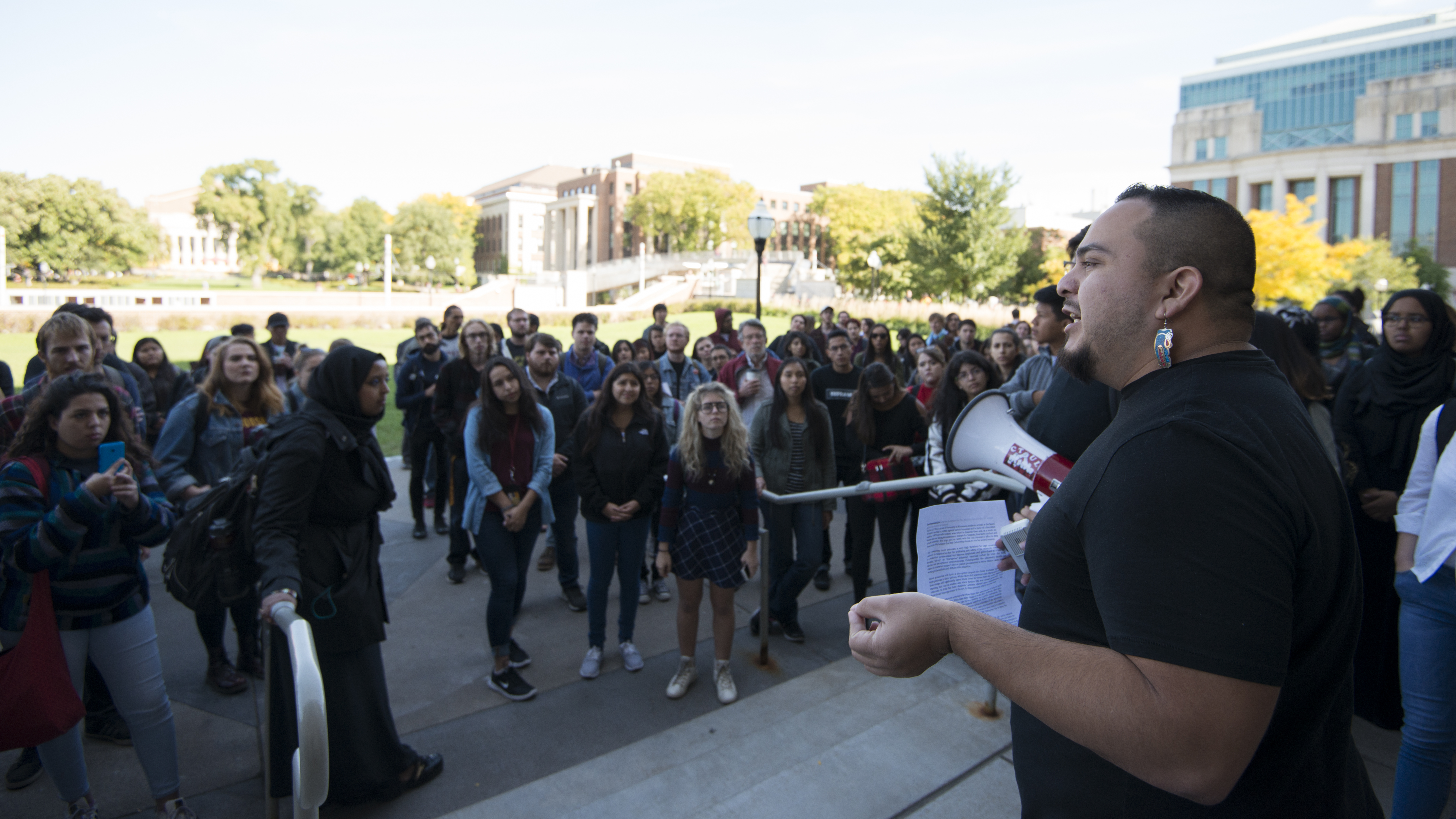 A man in a T-shirt stands at the head of a flight of steps with megaphone in hand, speaking to a small group of students.