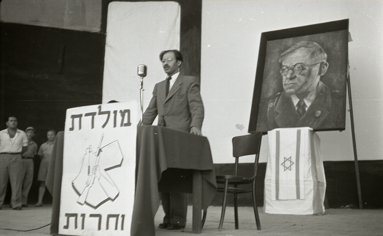 Menachem Begin stands at a podium in front of a map with borders that include large portions of modern-day Transjordan, Syria, and other countries.