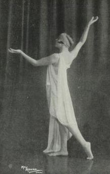 A young white woman wearing loose white draped cloths and a headwrap, barefoot, posed in front of a curtain.