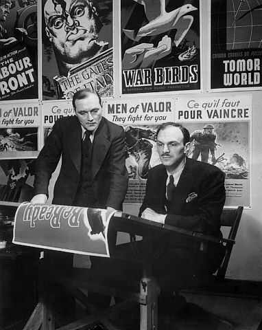 John Grierson (left), Chairman of the Wartime Information Board, meeting with Ralph Foster, Head of Graphics, National Film Board of Canada, to examine a series of posters produced by the National Film Board of Canada