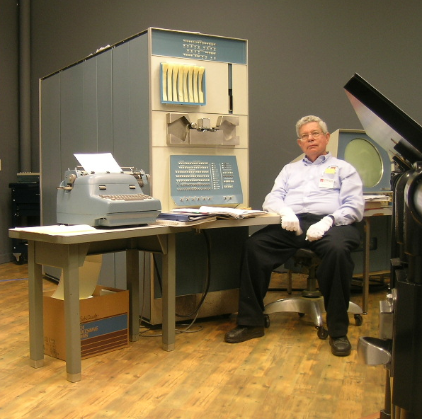 PDP-1 i Steve Russell, twórca gry Spacewar!. Pośrodku, nad biurkiem, główny panel sterowania, nad nim (srebrny) czytnik taśm perforowanych, pionowy slot powyżej to wyjście taśmy z dalekopisu, a obok podajnik na osiem taśm perforowanych.