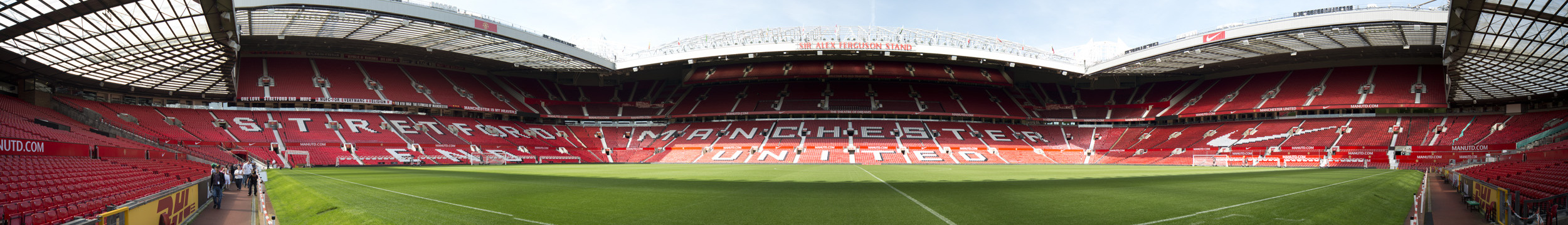 Uma arquibancada de um estádio de futebol.  Os assentos são vermelhos e as palavras "Manchester United" estão escritas em assentos brancos.  A cobertura do estande é sustentada por uma estrutura em balanço.  Na borda do telhado, está escrito "Old Trafford Manchester".