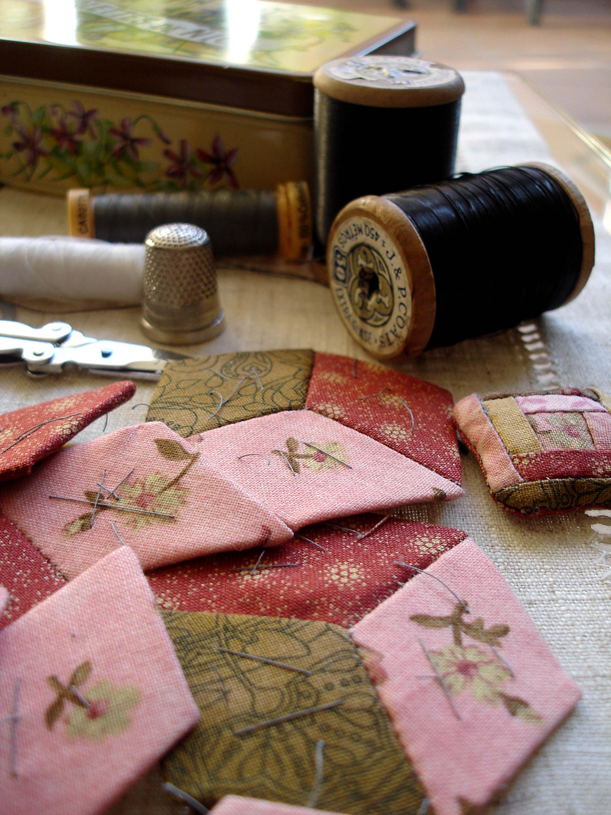 Diamonds of fabric tacked to paper have been pieced in threes into hexagons. In the background are scissors, reels of thread, a thimble, and a tin.