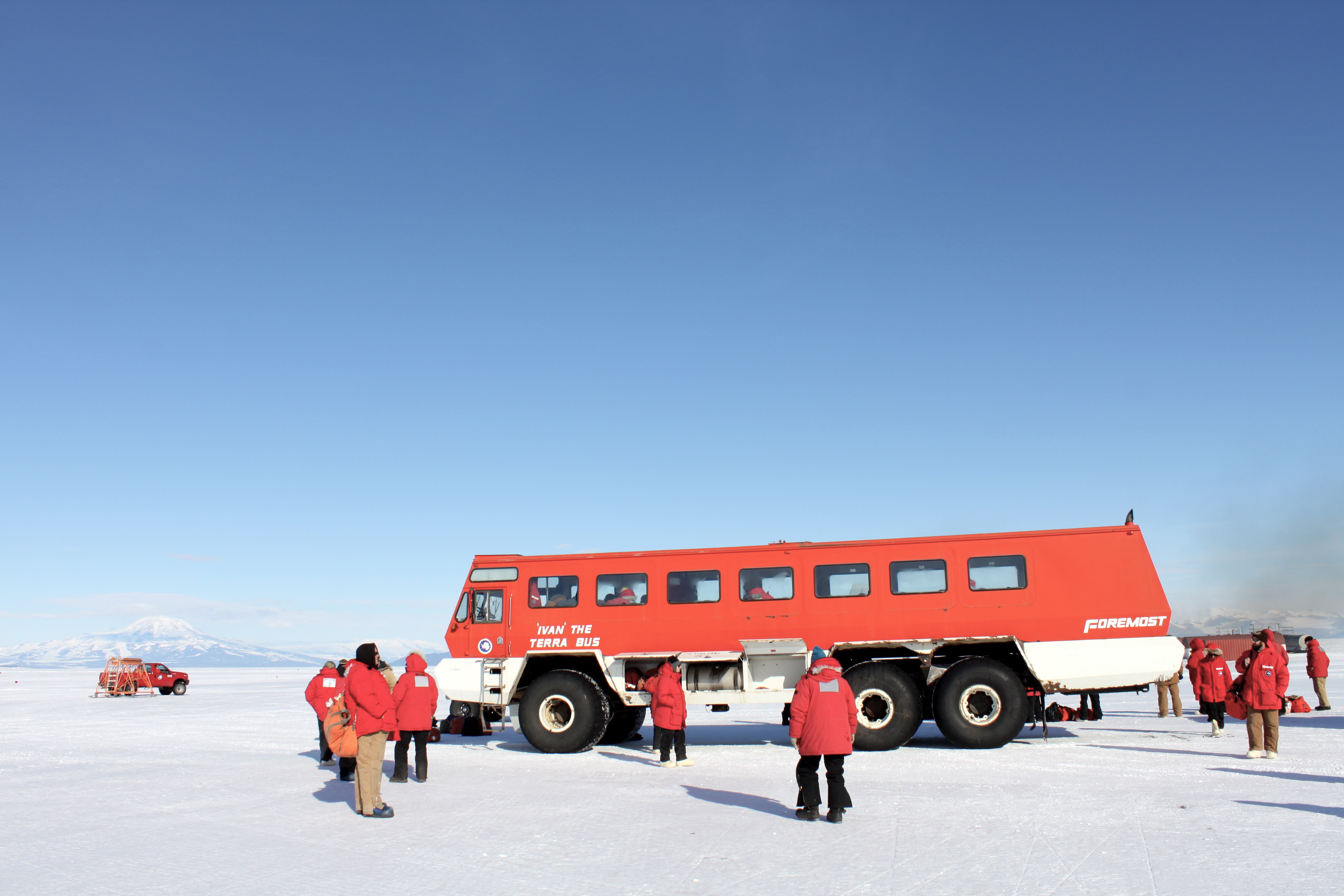 An image of Ivan the Terra Bus in Antarctica. Blue sky, white snow, and a few people standing around in red coats.