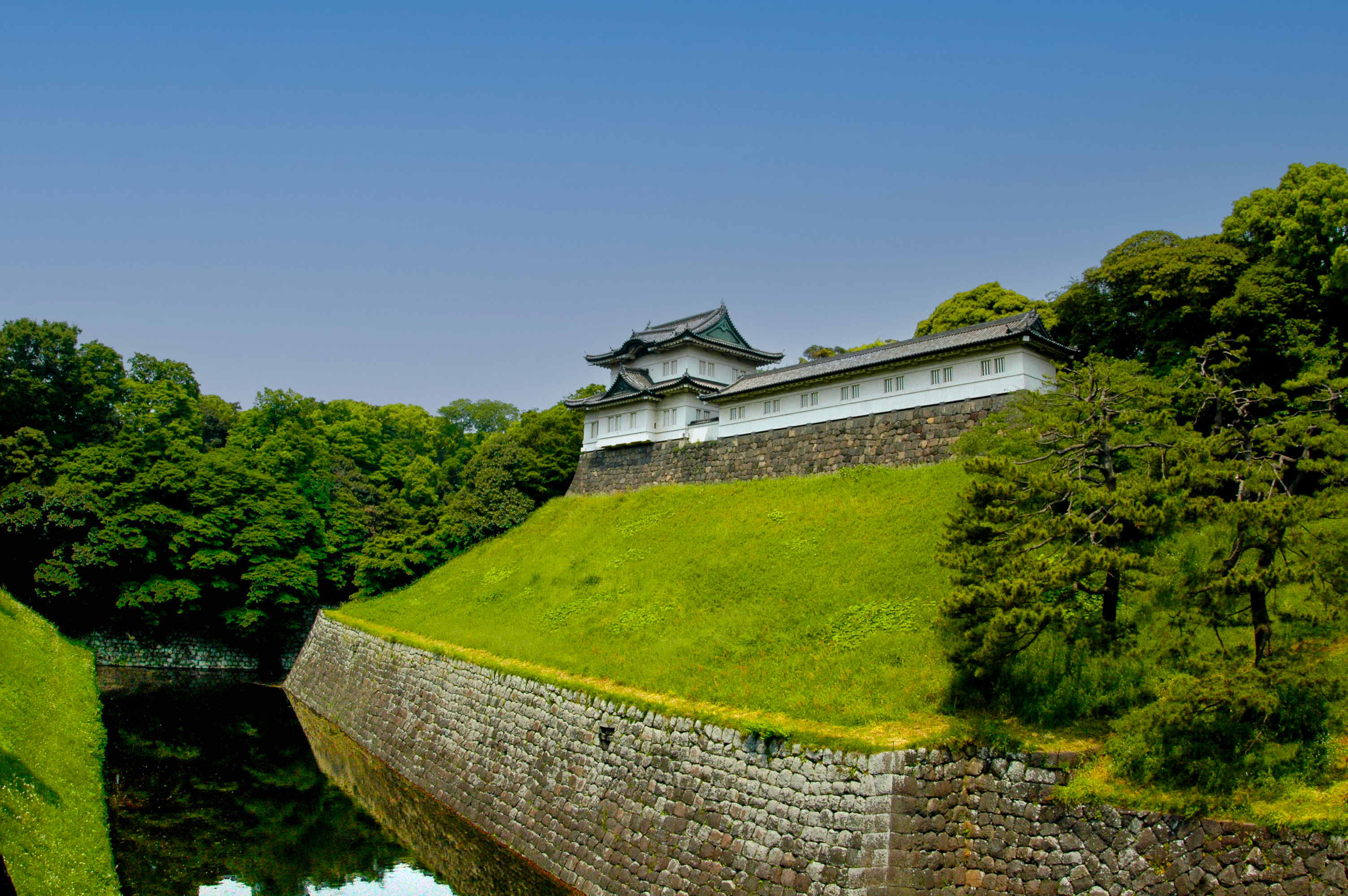 A photograph showing the keep of the Edo Castle on top of a grassy hill from the back, with a waterway below the hill. The hill is held in by a retaining wall.