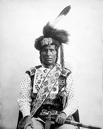Midwewinind ("Hij die van een afstand wordt geroepen") van Gaa-waabaabiganikaag (White Earth), Minnesota, 1894. Zijn kleding is typerend voor de zuidelijke Ojibweg in de 19e eeuw: met kralen bestikte zwarte stof en geborduurde sjerp met bloemmotieven en een tulband van bont. Op zijn tulband prijkt een arendsveer. In zijn hand draagt hij een bagamaagan of tomahawk-pijp.