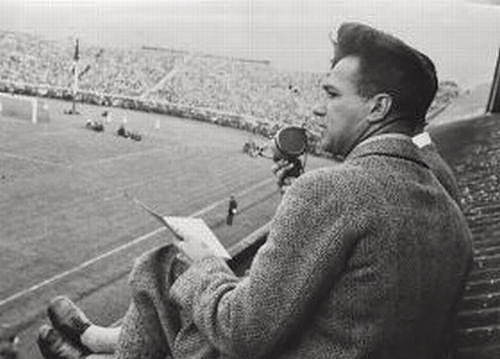 alt=Journalist, seated in the stands and speaking into a microphone