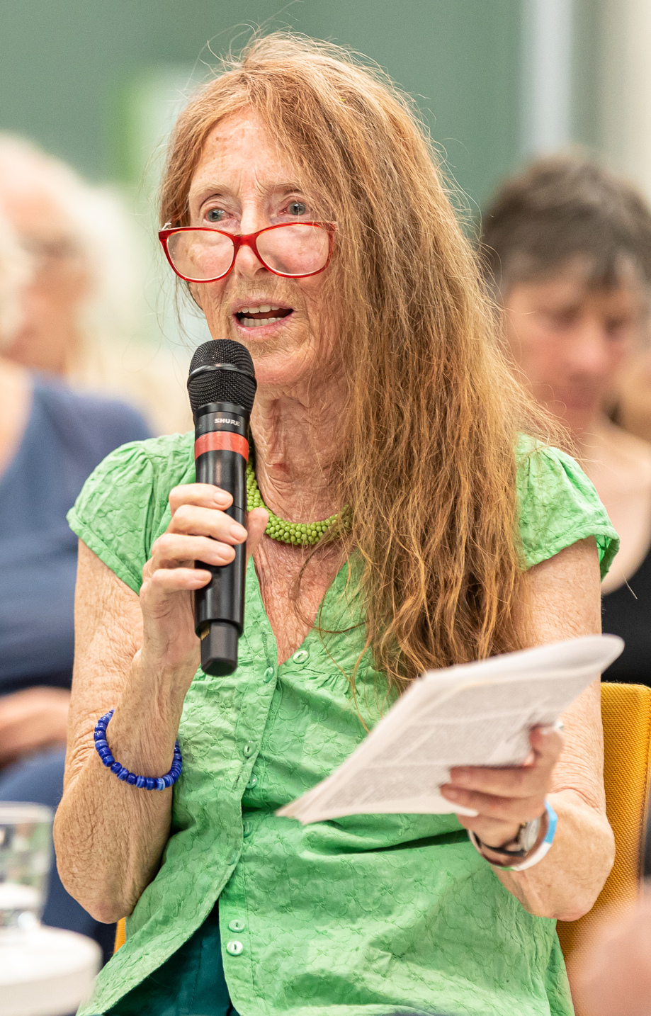 alternativtext=Eva Quistorp sitzt. Sie spricht in ein Mikrofon, das sie in der rechten Hand hält. In der linken Hand hält sie ein Schriftzug. Eva Quistorp hat schulterlanges rotes Haar, sie trägt eine rote Brille und eine grüne Bluse.