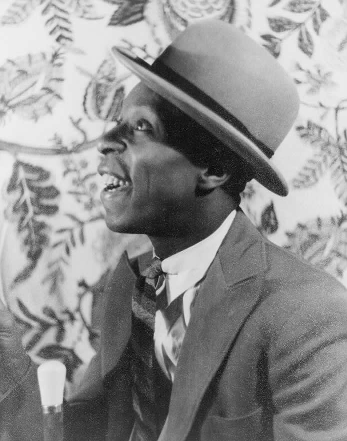 Left profile of African-American man in suit, bowler hat, and tie, with open-mouthed grin.