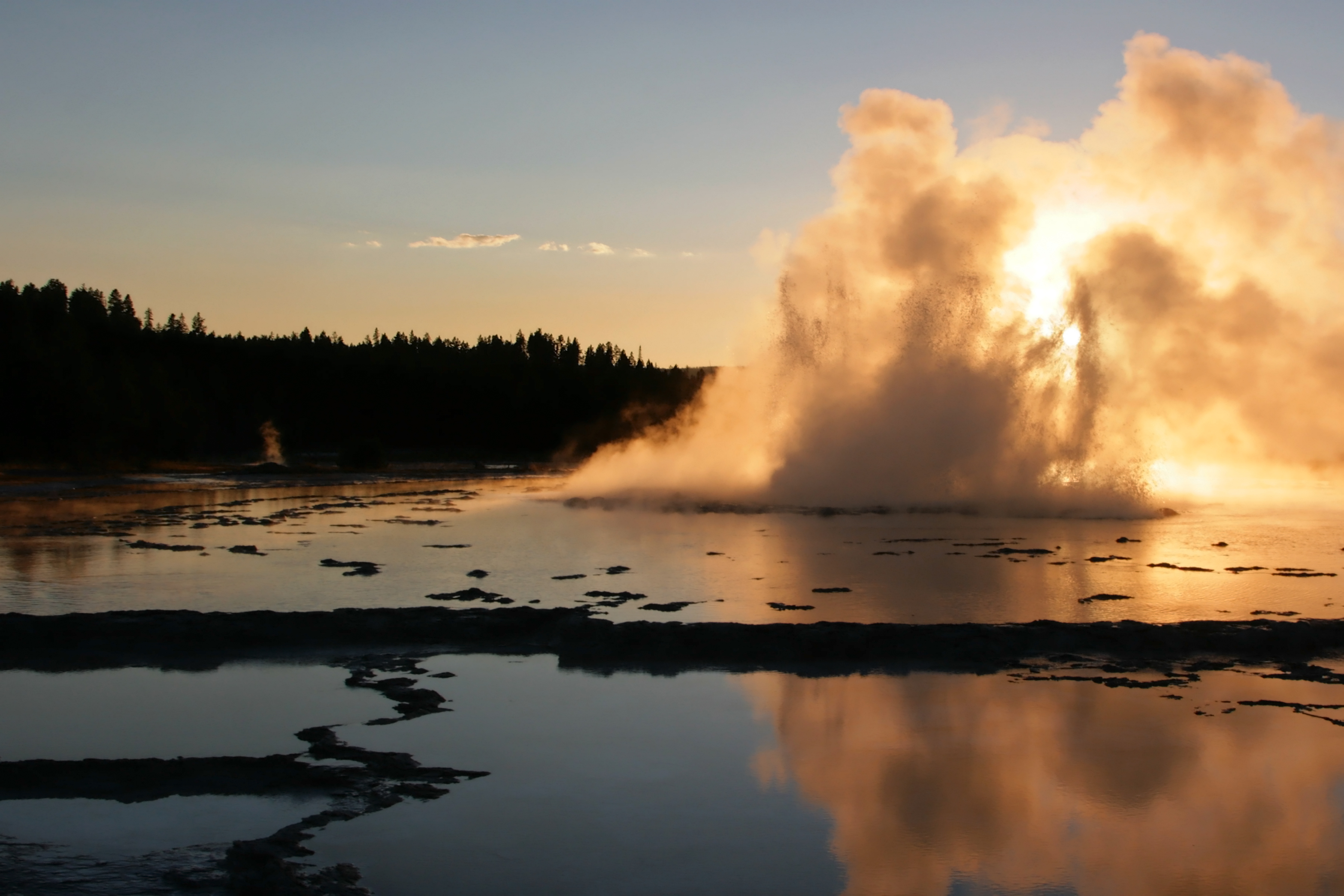 a horizontal picture of great fountain geyser at sunset. on the right is a picture of a geyser bursting into the sky, illuminated by the sunset. on the left are a line of trees blackened by shadow. at the bottom is a lake reflecting the geyser, sky, and tree line. 