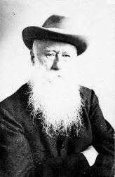 A black and white photograph of a man from the chest up. He is wearing a hat and sports a long white beard. His arms are crossed.