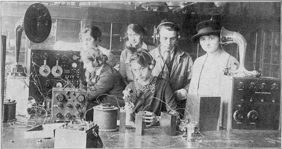 A group of students sit around radio equipment in a black-and-white photo from the 1920s, sitting at a table covered with wires and radio equipment.
