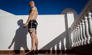 GIF animado de homem usando bermuda preta e sem camisa, pele clara, realizando os movimentos do exercício burpee em área externa, em frente a um muro e sacada.