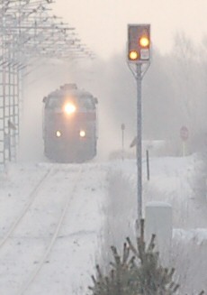 A Finnish distant signal at the western approach to Muhos station is displaying Expect Stop. In the background, express train 81 is pulling away from the station.