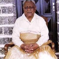 Photo en couleur d'un homme âgé ceint d'une natte traditionnelle (ta'ovala) et vêtu d'une chemise blanche, assis sur un trône en bois, devant une grande porte en bois et un mur en pierres apparentes.