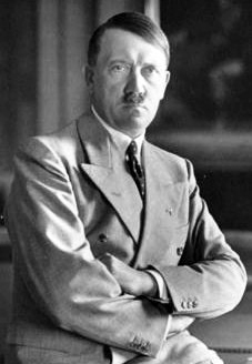 A black and white photo of a man leaning against a desk staring into the camera. His arms are crossed and he is wearing a tie underneath his overcoat. An office can be seen in the background of the photo.