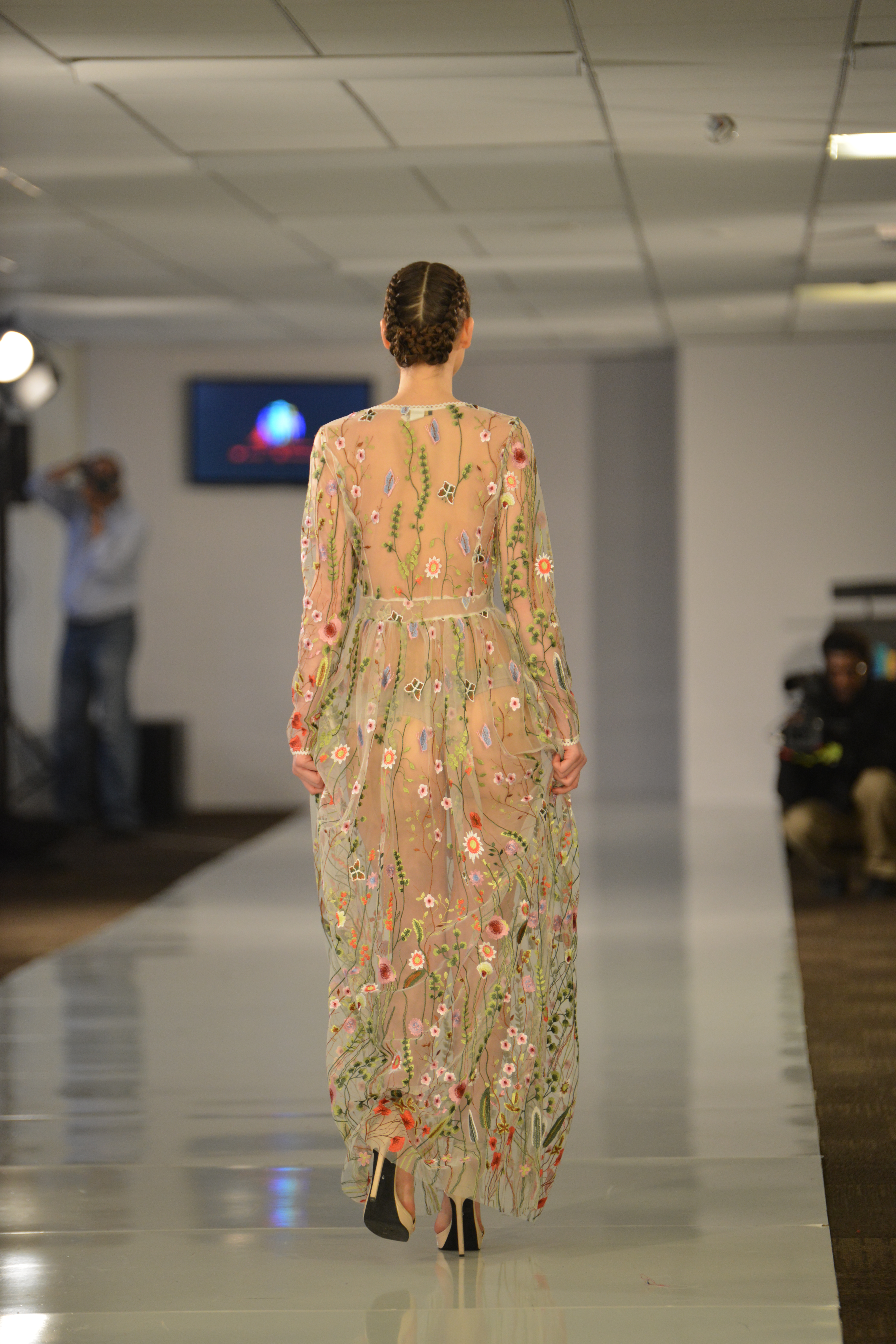 A woman in a long-sleeved full-length sheer embroidered dress walks away down a catwalk.