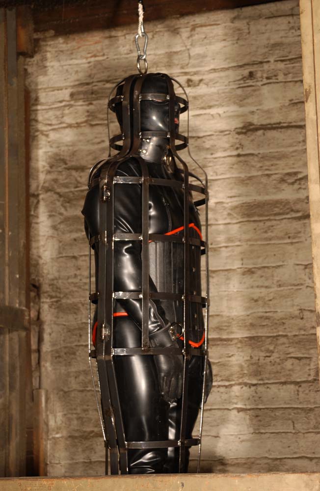 Subject wearing a latex outfit and held captive in a suspension cage.jpg. 