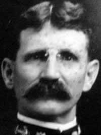 Foto frontal en blanco y negro de un hombre con gafas pequeñas y un bigote cuidadosamente recortado.  El borde de un uniforme de la Marina está alrededor de su cuello.