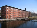 A five-story oblong brick building (5 window bays on the short side, 35 on the long side) stands beside the Charles River. A portion of a wooden dam is visible to the right of the building.