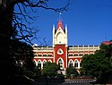 A red and white building with a central tower topped by a flag all under a clear blue sky and with green trees in the foreground