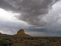 Тамни, котрљајући олујни облаци се спуштају изнад пустињског пејзажа; стрмо брдо стоји у непосредној близини, лево од центра.