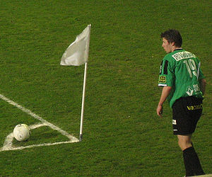 A man wearing a green top and black shorts, standing on a grass field. In front of him is a white flag, on a flagpole that appears to be slighly shorter than the man. On the ground next to the flagpole is a football.