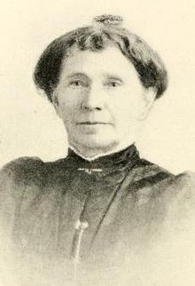 An older white woman wearing a dark high-collared dress.