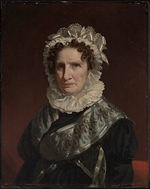 Portrait of an older woman, at half length, against a plain background. She wears a lace bonnet and ruff, and black dress with silver-gray sash and trim. Her hair is curly and auburn. She has a very pale complexion.