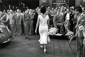 Young actress and circus performer Moira Orfei walks towards the Galleria Vittorio Emanuele II in Milan, while a large group of men turn to look at her. 1954