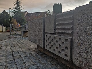 The concrete fountain which today is located at the intersection of Zihlstrasse and Gstaldenstrasse in Hinwil was designed by Max Vogt as part of the Hinwil railway station. It has since been moved away from the railway station and placed here. This photo is a close-up of the fountain's outer wall which is facing Gstaldenstrasse and features an interpretation of the Hinwil municipality's coat of arms using patterns to represent colours