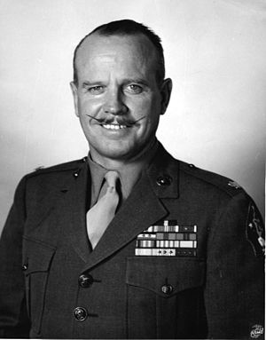 Head and torso of white man with slightly receding hairline and waxed mustache wearing tan shirt and necktie, green blouse with U.S. Marine lieutenant colonel's insignia, five rows of ribbons on his chest, and 2nd Marine Division patch on his left shoulder. The U.S. Marine Corps logo is superimposed on the lower right corner of this black-and-white photograph.